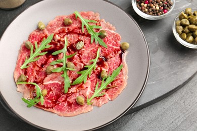 Photo of Fresh beef carpaccio with arugula, capers and sauce on dark textured table, above view