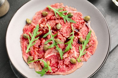 Photo of Fresh beef carpaccio with arugula, capers and sauce on dark textured table, top view