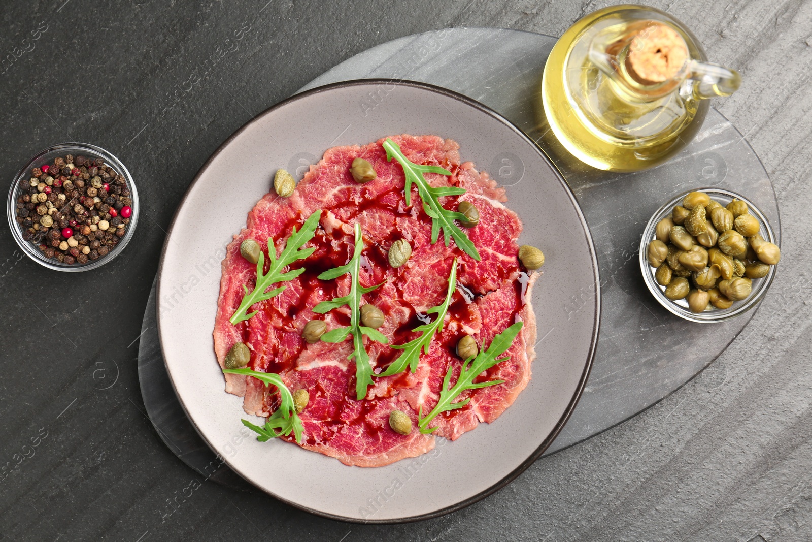 Photo of Fresh beef carpaccio with arugula, capers and sauce on dark textured table, flat lay