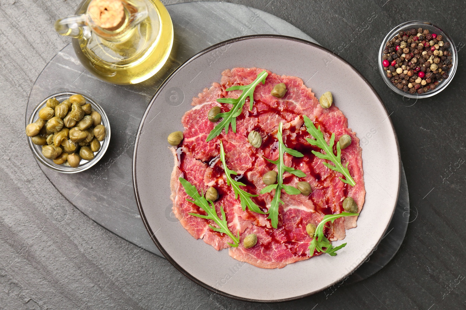 Photo of Fresh beef carpaccio with arugula, capers and sauce on dark textured table, flat lay