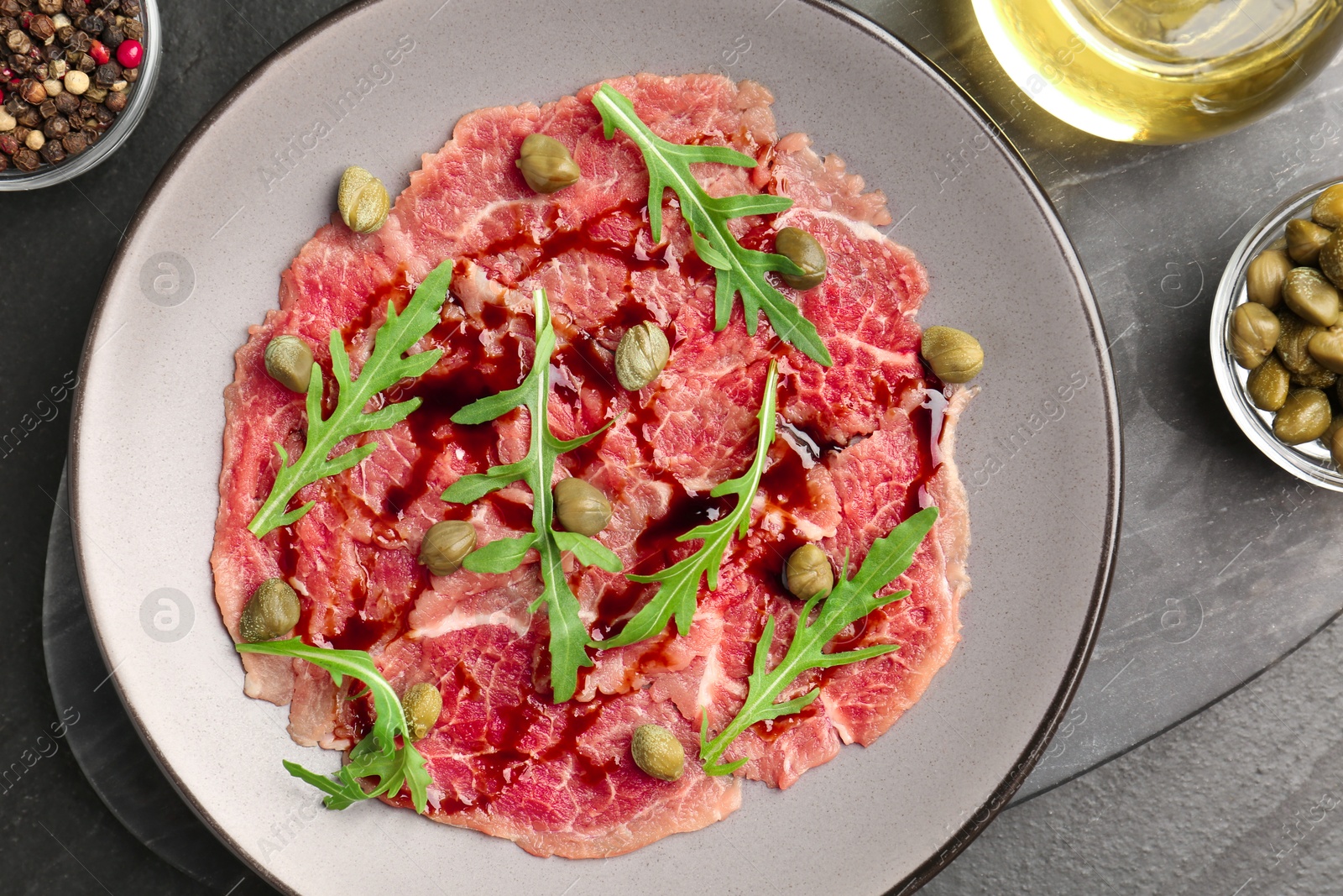 Photo of Fresh beef carpaccio with arugula, capers and sauce on dark textured table, flat lay