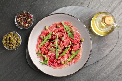 Photo of Fresh beef carpaccio with arugula, capers and sauce on dark textured table, flat lay