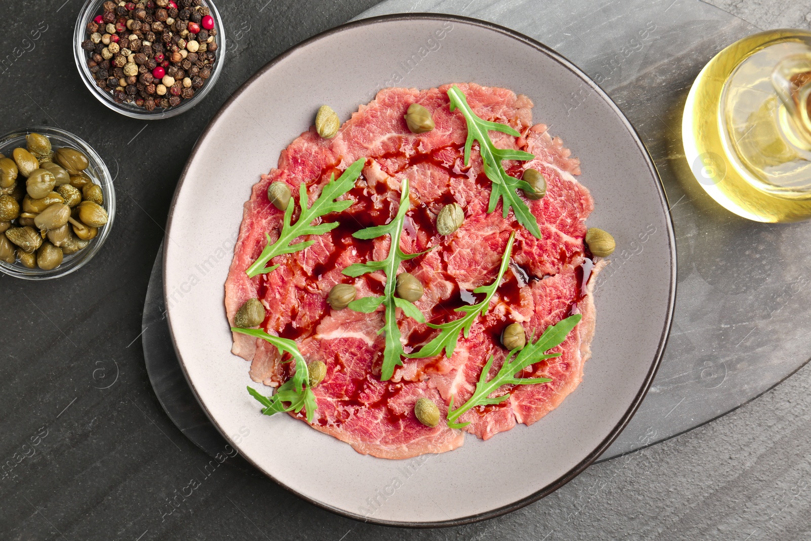 Photo of Fresh beef carpaccio with arugula, capers and sauce on dark textured table, flat lay