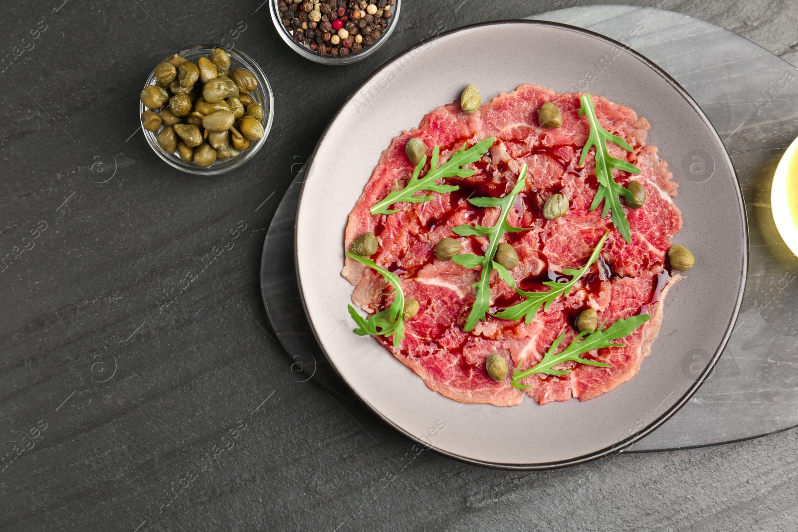 Photo of Fresh beef carpaccio with arugula, capers and sauce on dark textured table, flat lay. Space for text