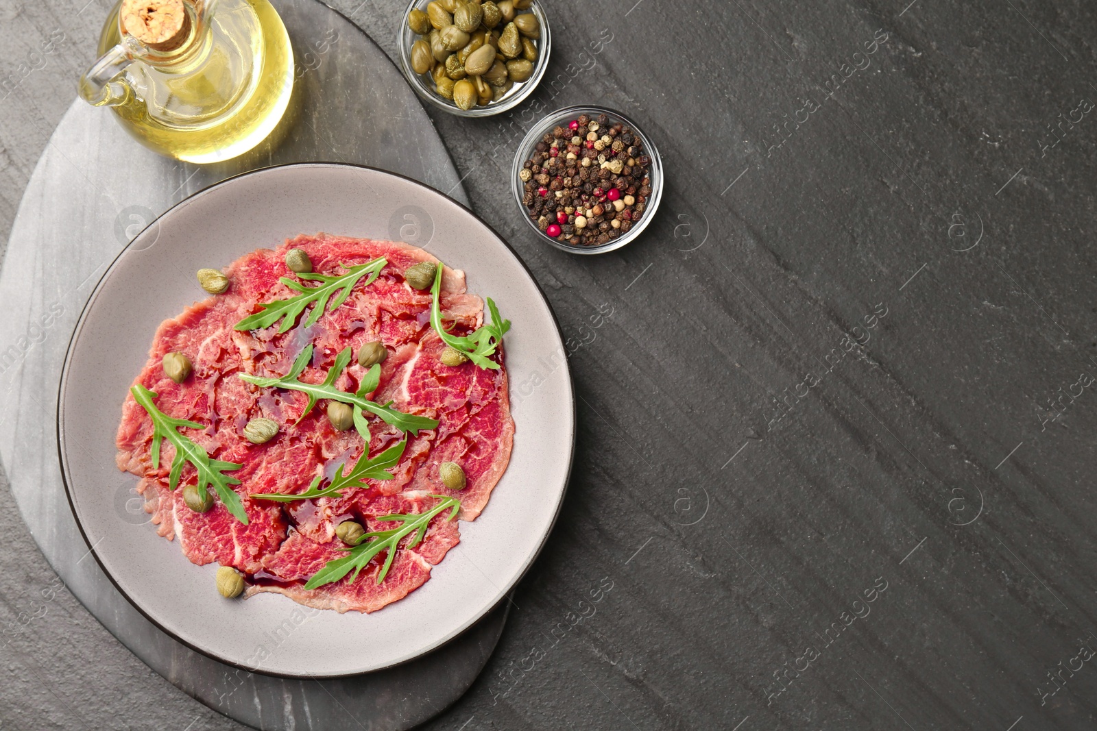 Photo of Fresh beef carpaccio with arugula, capers and sauce on dark textured table, flat lay. Space for text