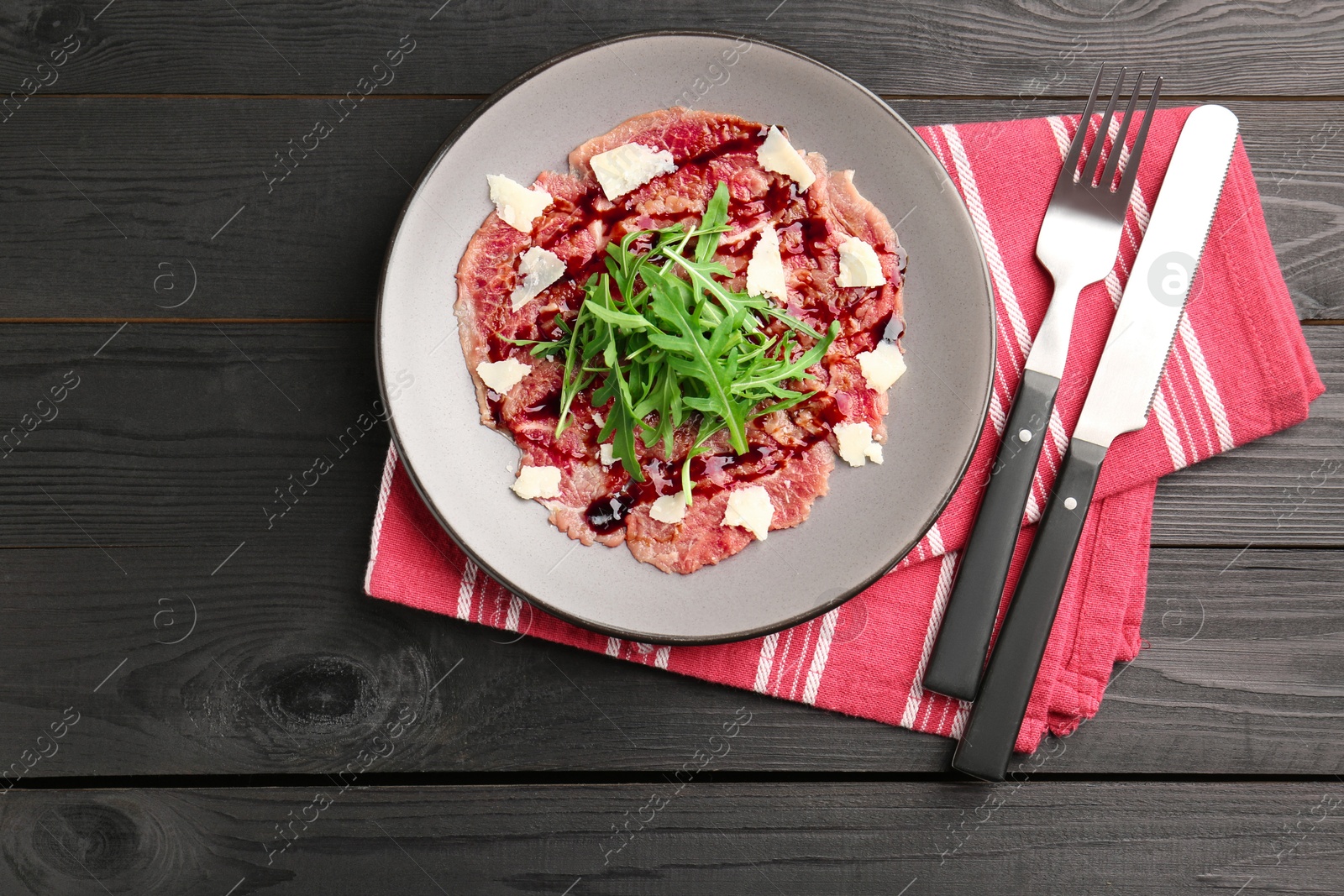 Photo of Fresh beef carpaccio with arugula, cheese and sauce on black wooden table, top view