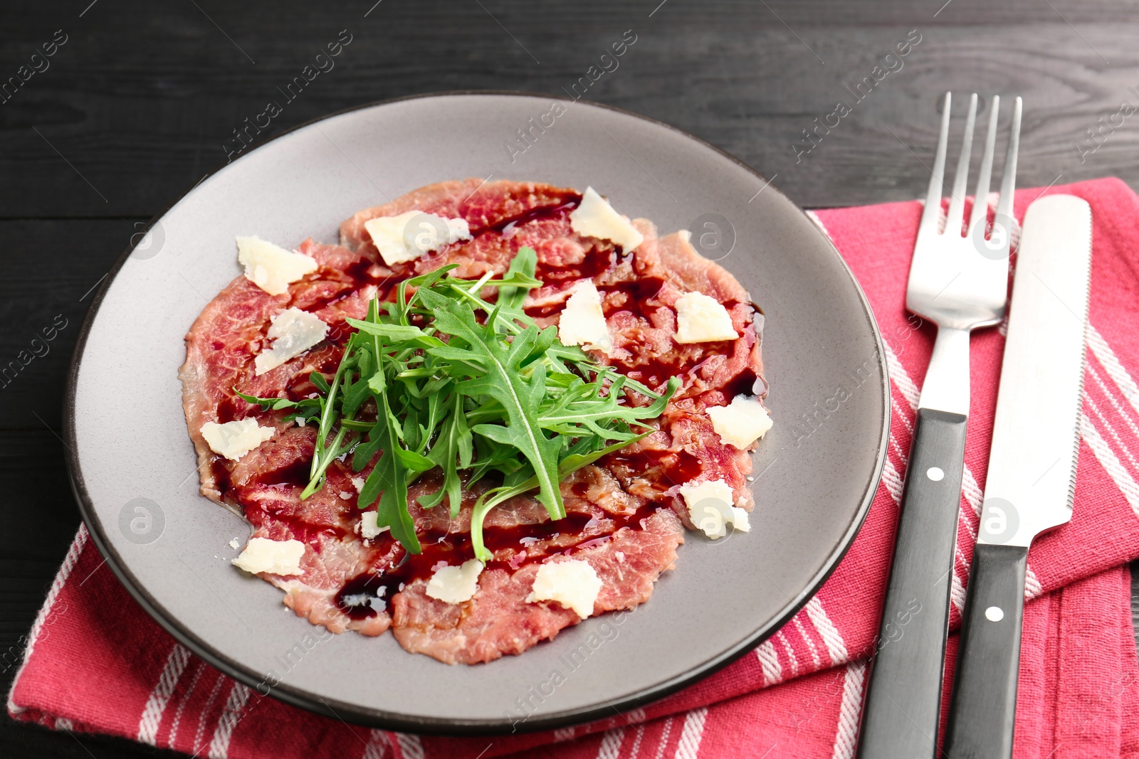 Photo of Fresh beef carpaccio with arugula, cheese and sauce on black wooden table, closeup