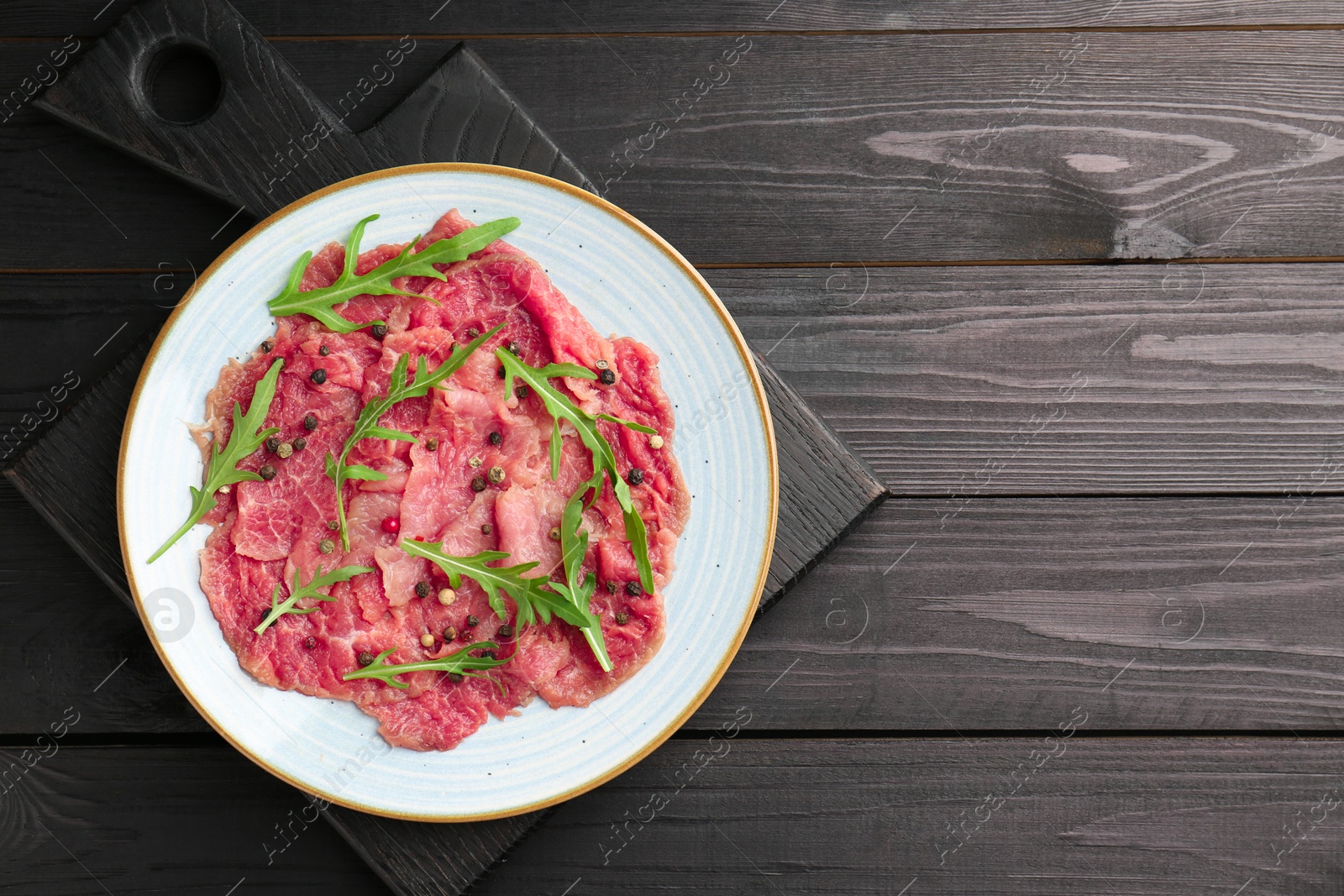 Photo of Fresh beef carpaccio with arugula and peppercorns on black wooden table, top view. Space for text