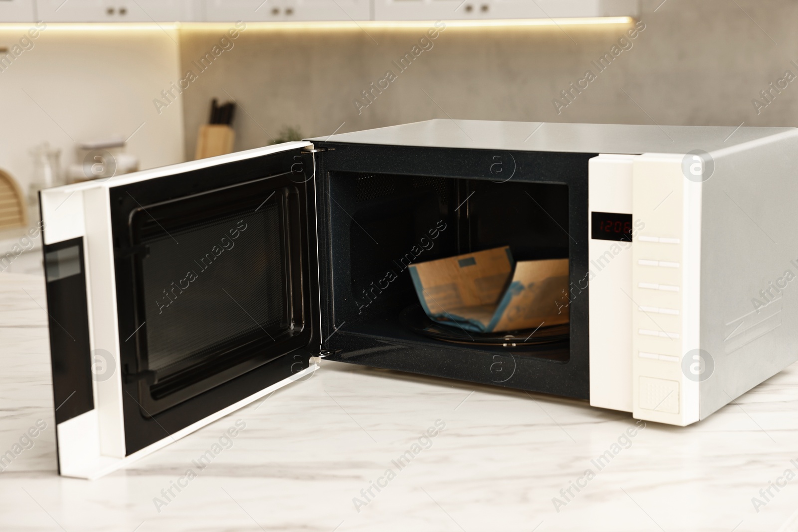 Photo of Open microwave oven with bag of popcorn on white marble table in kitchen