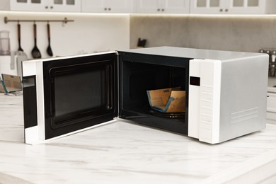 Photo of Open microwave oven with bag of popcorn on white marble table in kitchen