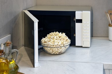 Photo of Tasty popcorn in bowl near microwave oven on white marble countertop indoors