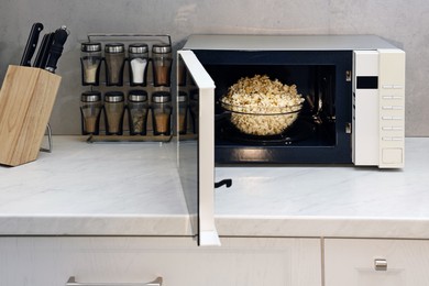 Photo of Open microwave oven with bowl of popcorn on white marble countertop in kitchen