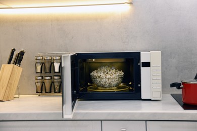 Photo of Open microwave oven with bowl of popcorn on white marble countertop in kitchen