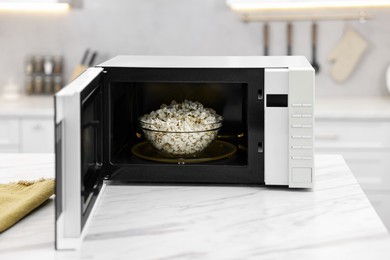 Photo of Open microwave oven with bowl of popcorn on white marble table in kitchen