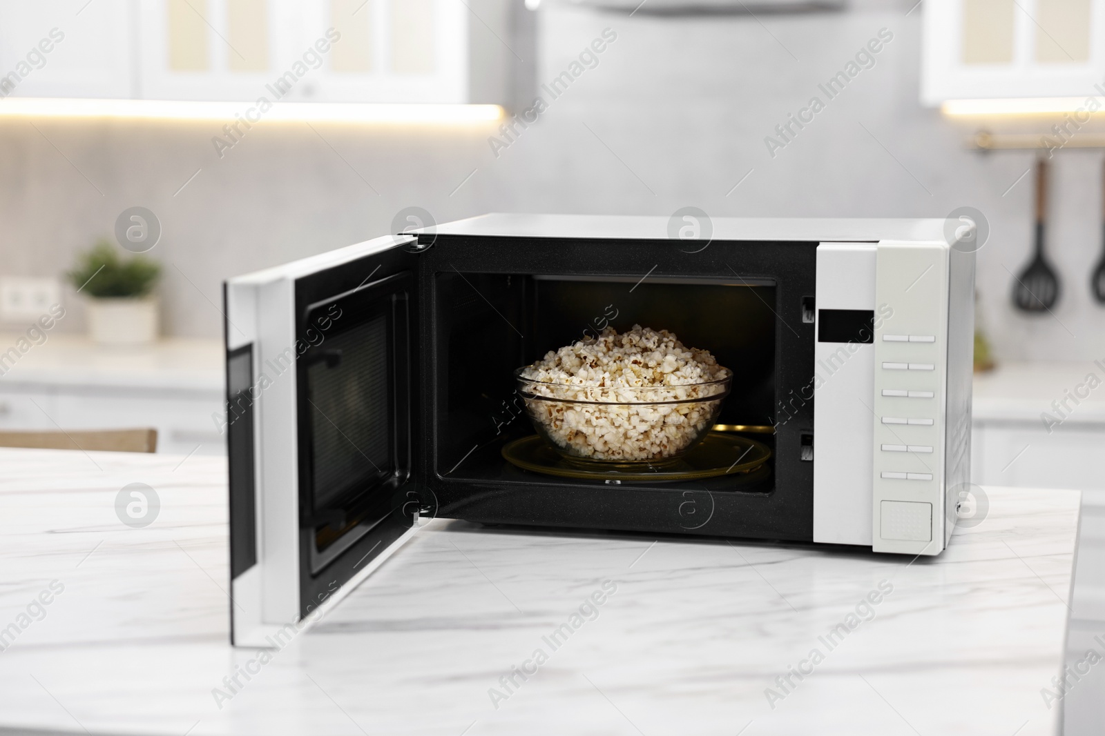 Photo of Open microwave oven with bowl of popcorn on white marble table in kitchen