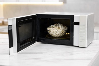 Photo of Open microwave oven with bowl of popcorn on white marble table in kitchen