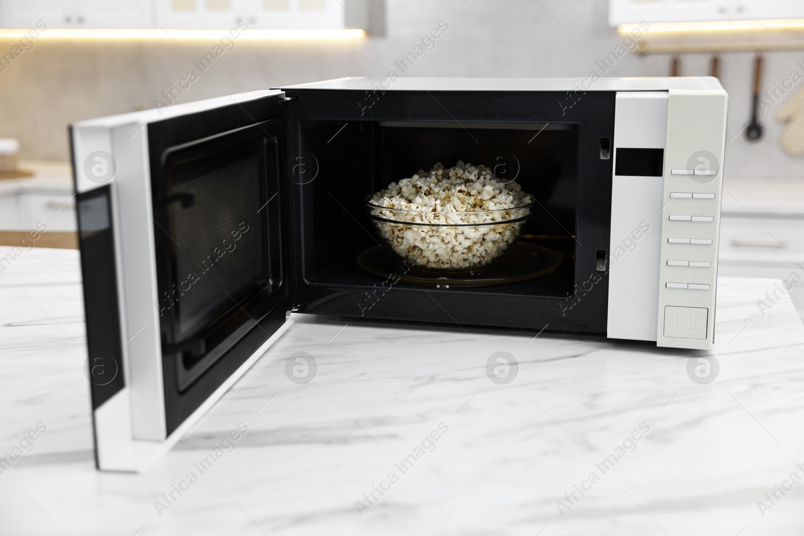 Photo of Open microwave oven with bowl of popcorn on white marble table in kitchen