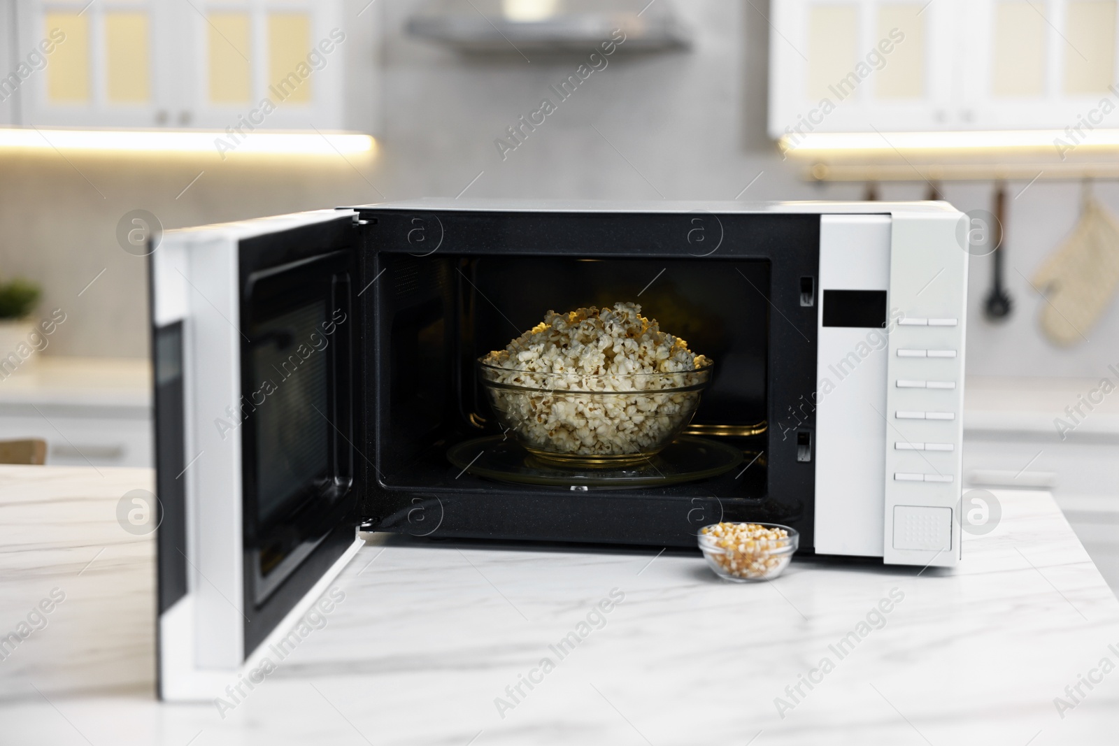 Photo of Open microwave oven with bowl of popcorn and kernels on white marble table in kitchen