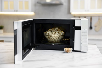 Photo of Open microwave oven with bowl of popcorn and kernels on white marble table in kitchen