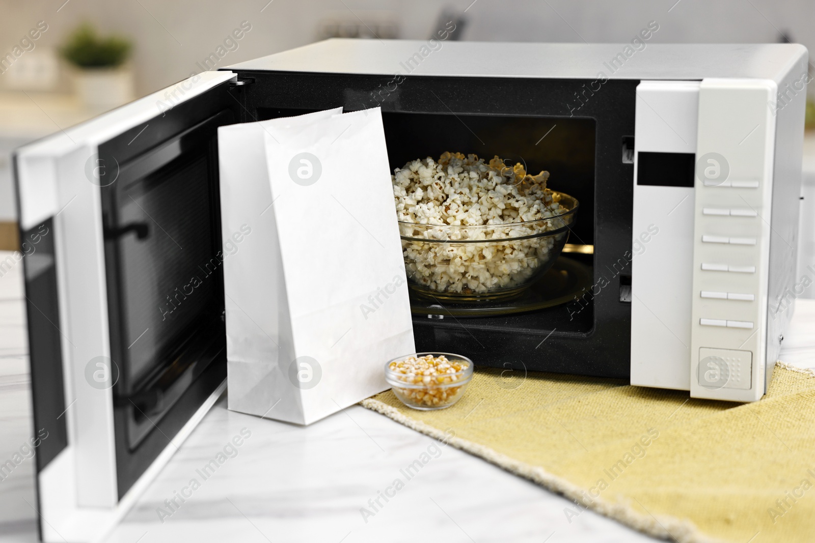 Photo of Open microwave oven with bowl of popcorn, paper bag and kernels on white marble table in kitchen