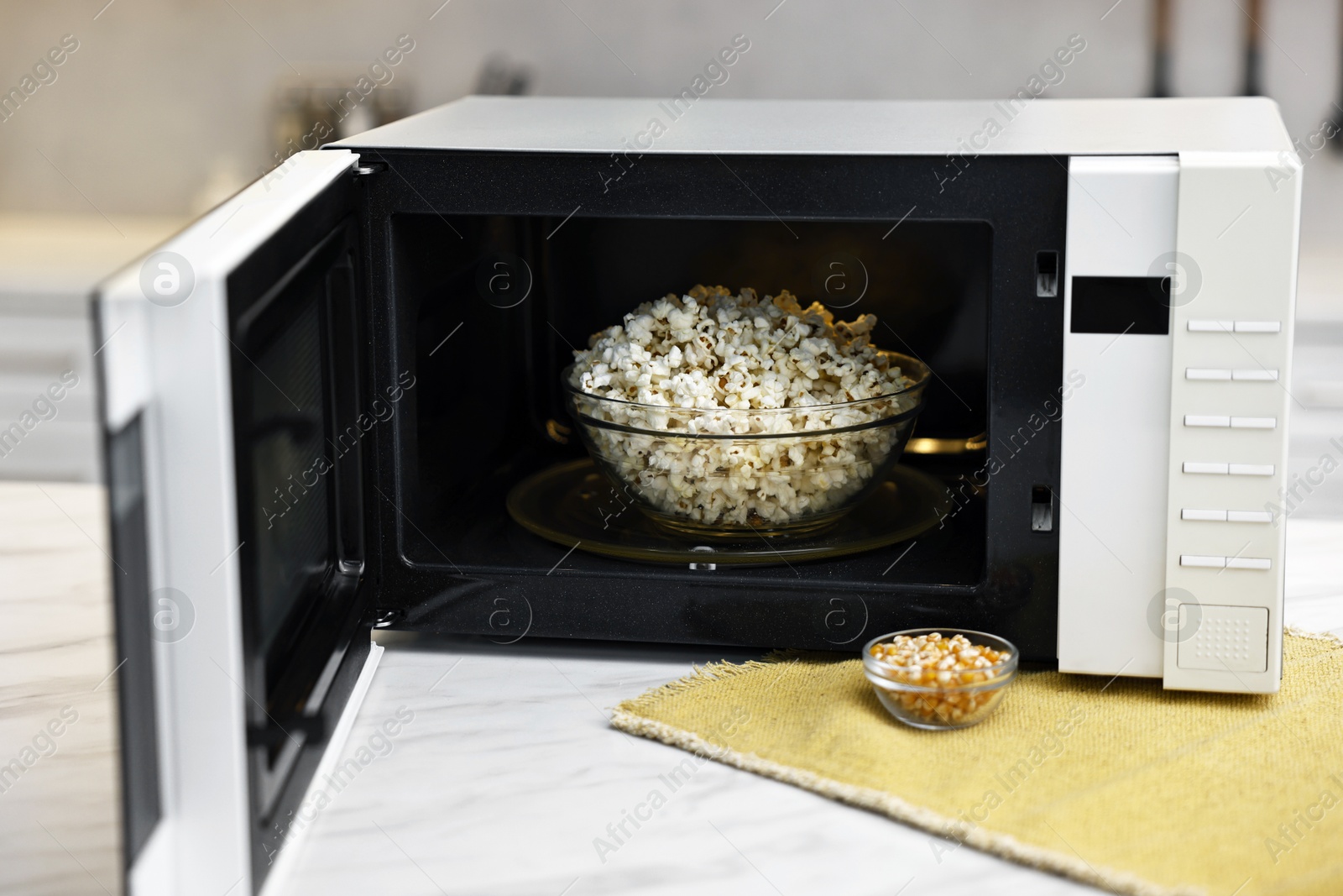 Photo of Open microwave oven with bowl of popcorn and kernels on white marble table in kitchen