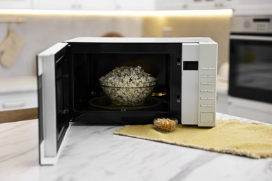 Photo of Open microwave oven with bowl of popcorn and kernels on white marble table in kitchen