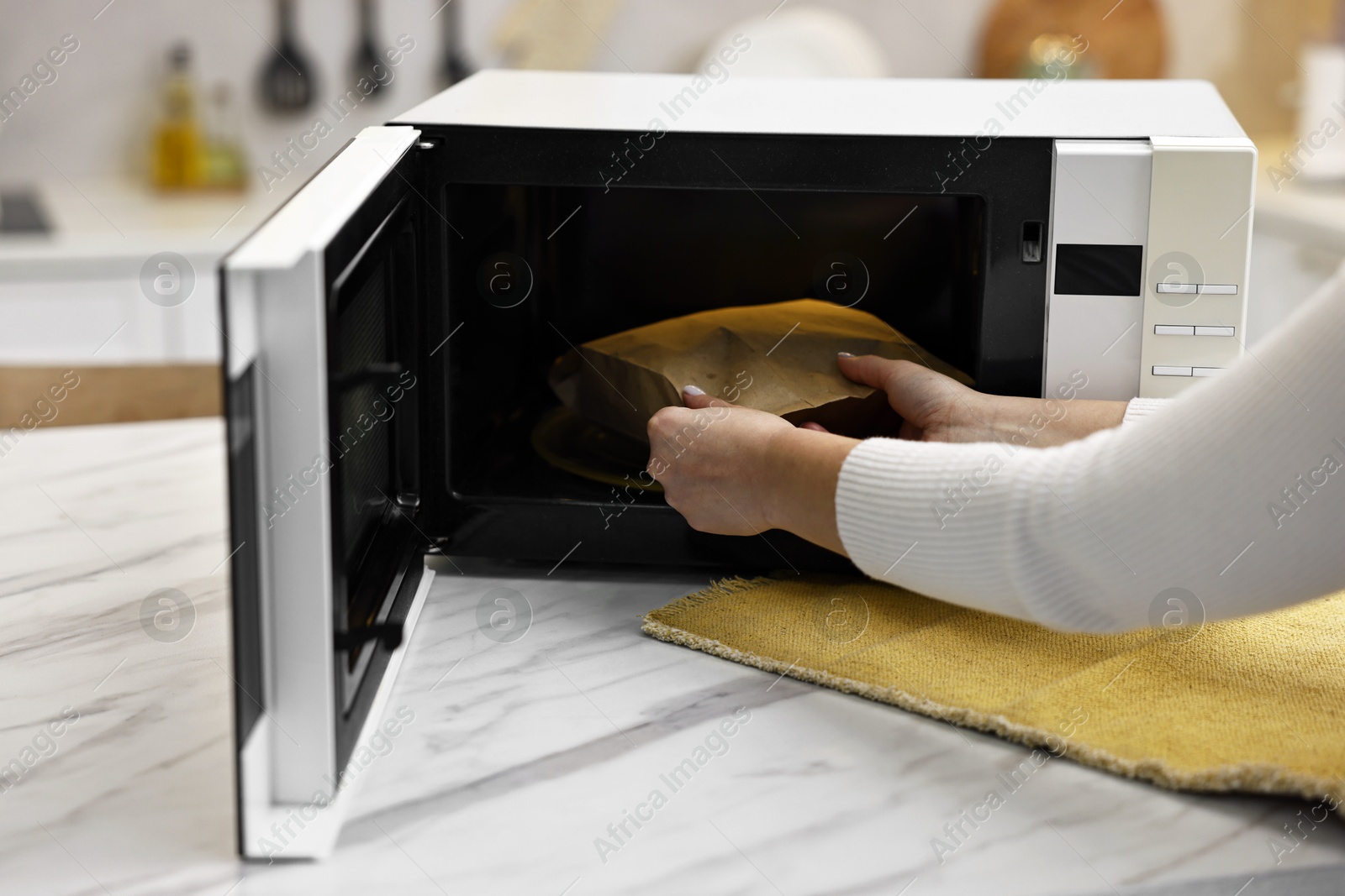 Photo of Woman taking bag with popcorn out of microwave oven at white marble table in kitchen, closeup