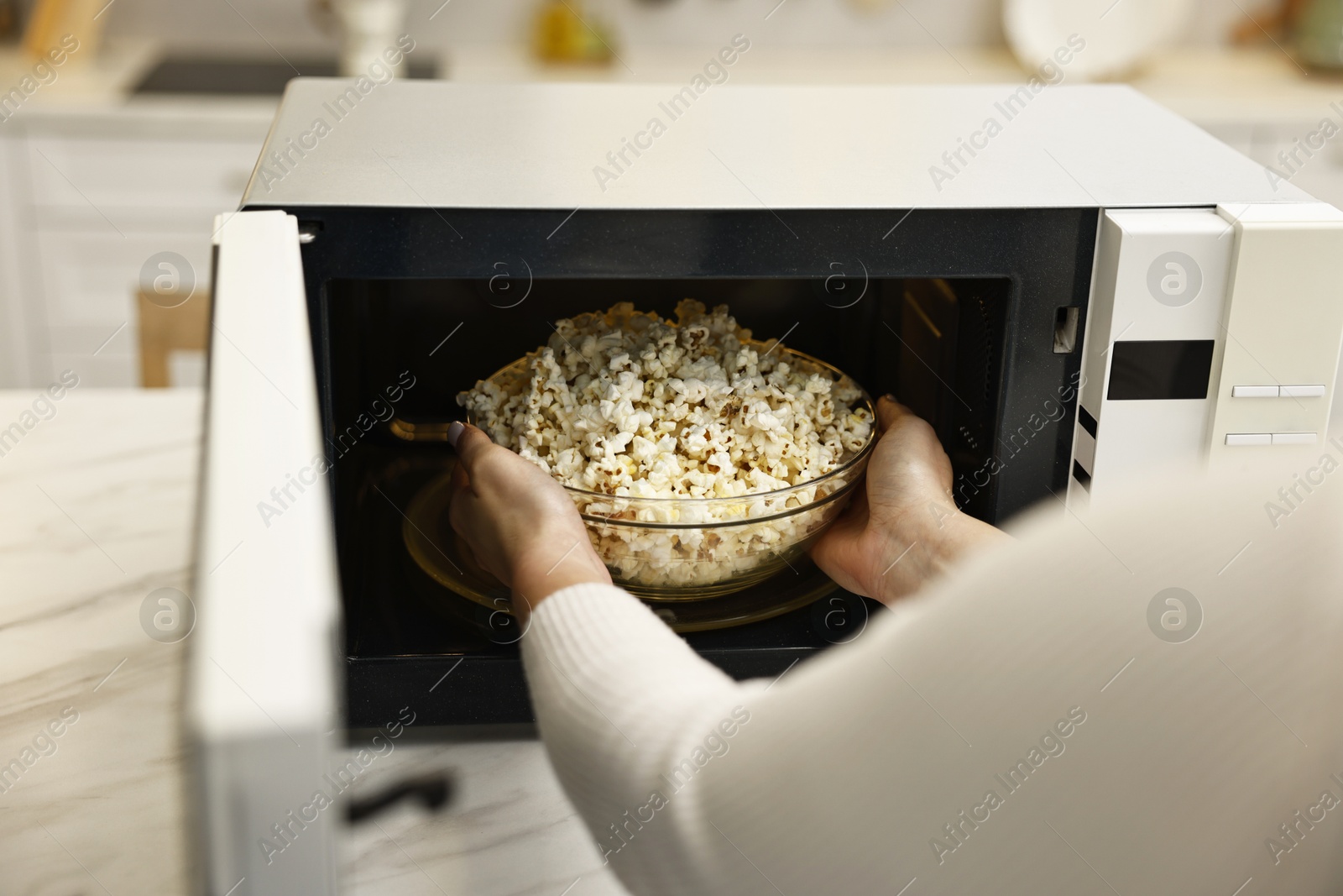 Photo of Woman taking bowl with popcorn out of microwave oven at white marble table in kitchen, closeup