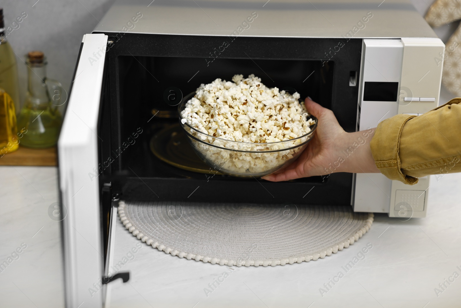 Photo of Woman taking bowl with popcorn out of microwave oven at white marble countertop in kitchen, closeup