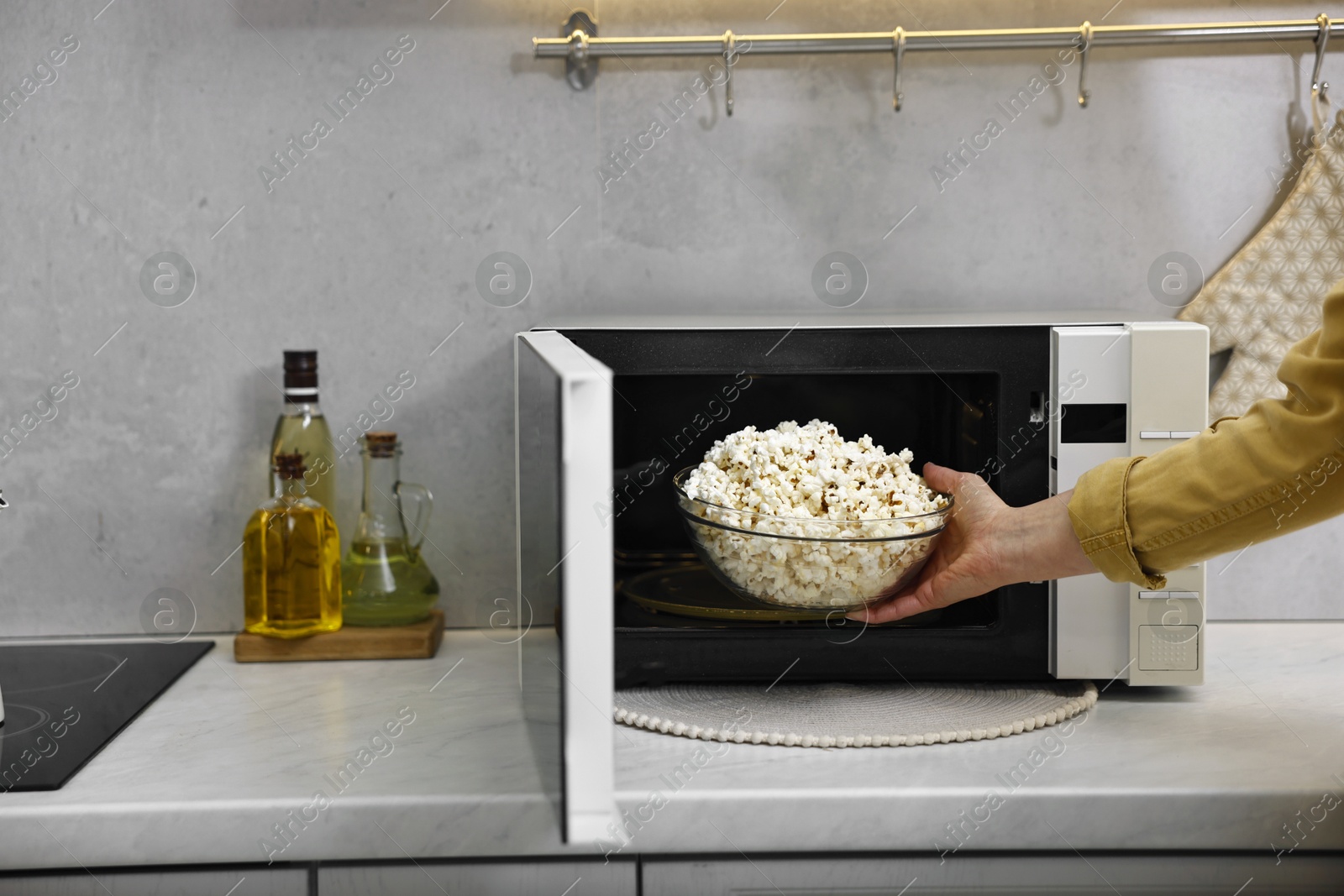 Photo of Woman taking bowl with popcorn out of microwave oven at white marble countertop in kitchen, closeup