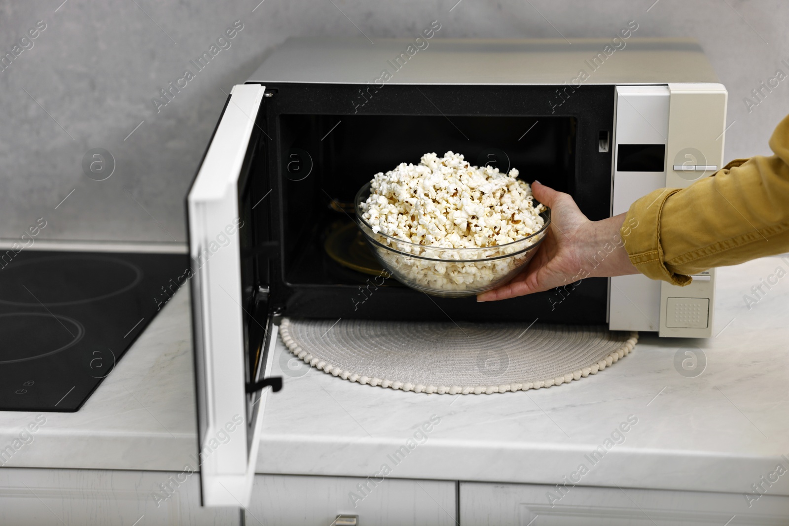 Photo of Woman taking bowl with popcorn out of microwave oven at white marble countertop in kitchen, closeup