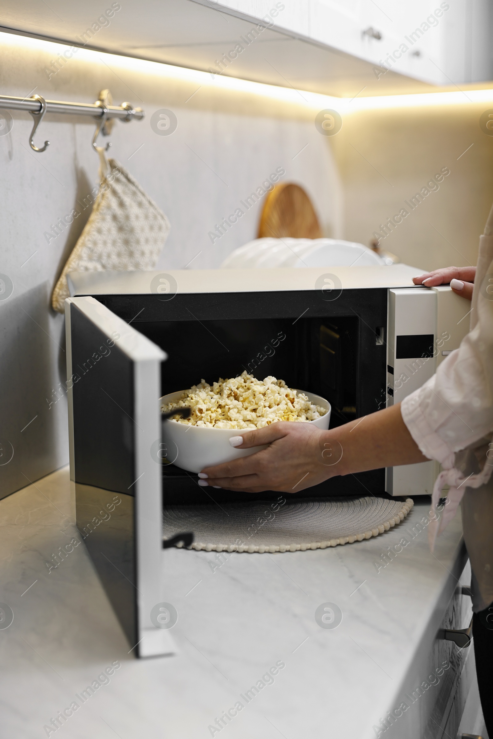 Photo of Woman taking bowl with popcorn out of microwave oven at white marble countertop in kitchen, closeup