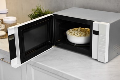 Photo of Open microwave oven with bowl of popcorn on white marble countertop in kitchen