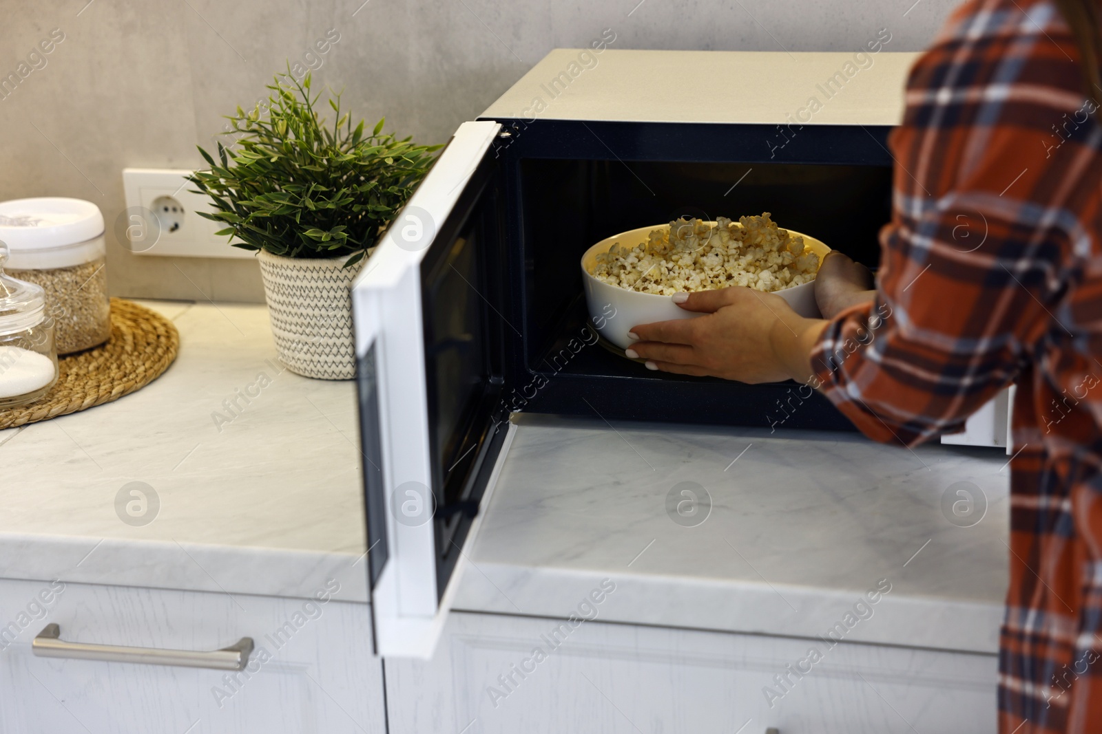 Photo of Woman taking bowl with popcorn out of microwave oven at white marble countertop in kitchen, closeup