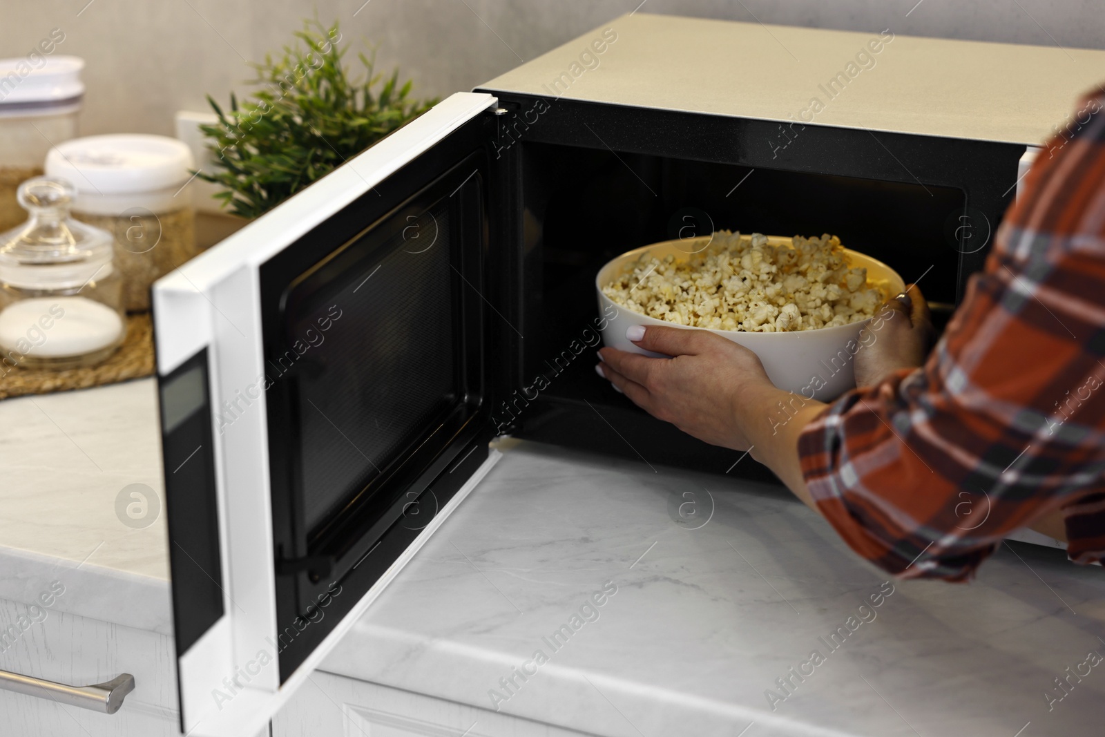 Photo of Woman taking bowl with popcorn out of microwave oven at white marble countertop in kitchen, closeup