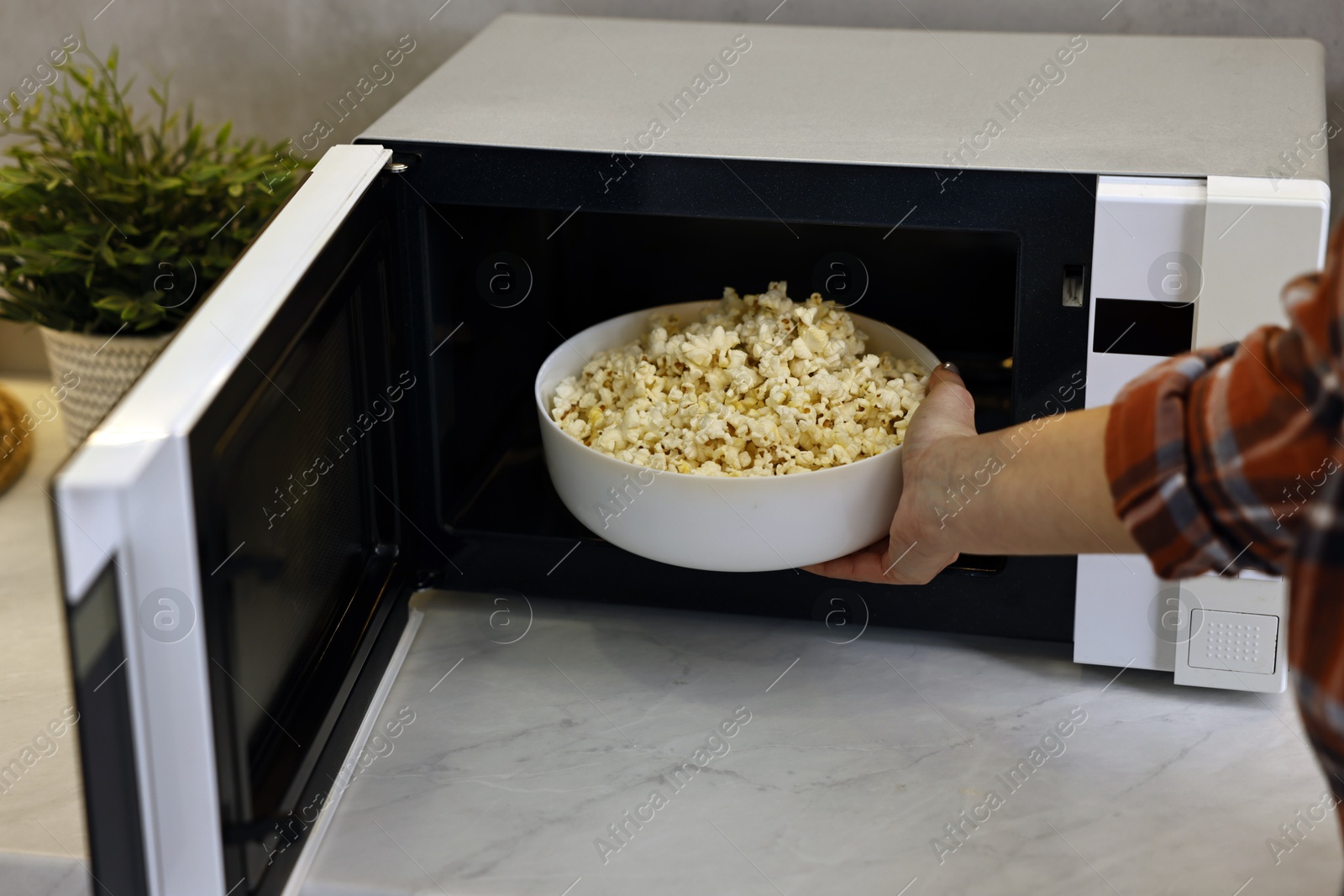 Photo of Woman taking bowl with popcorn out of microwave oven at white marble countertop in kitchen, closeup