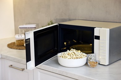 Photo of Open microwave oven with bowl of popcorn, kernels and bag on white marble countertop in kitchen