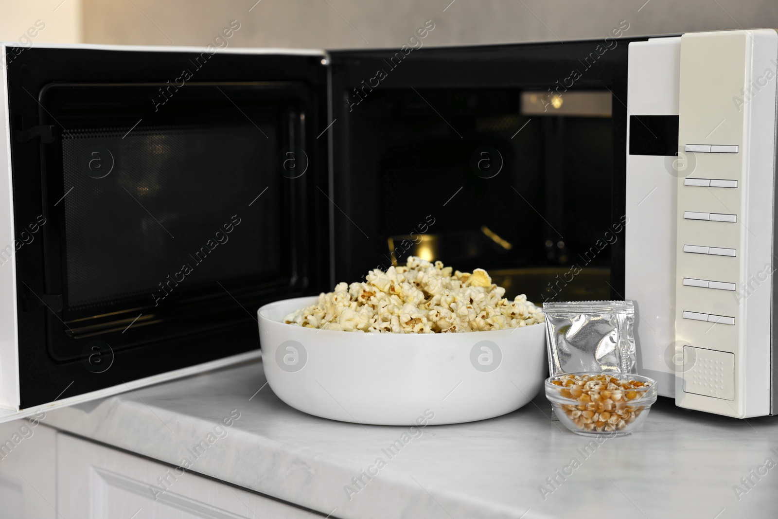 Photo of Open microwave oven with bowl of popcorn, kernels and bag on white marble countertop in kitchen