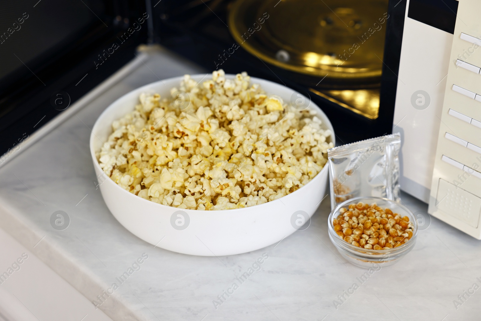 Photo of Bowl of popcorn, kernels and bag near open microwave oven on white marble countertop, closeup