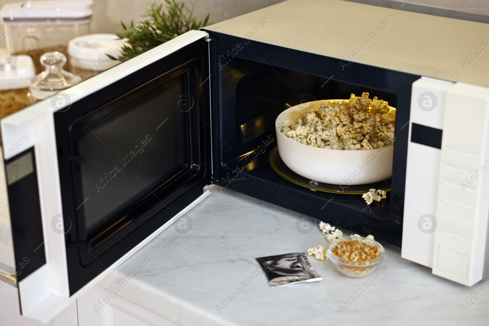 Photo of Open microwave oven with bowl of popcorn, kernels and bag on white marble countertop in kitchen