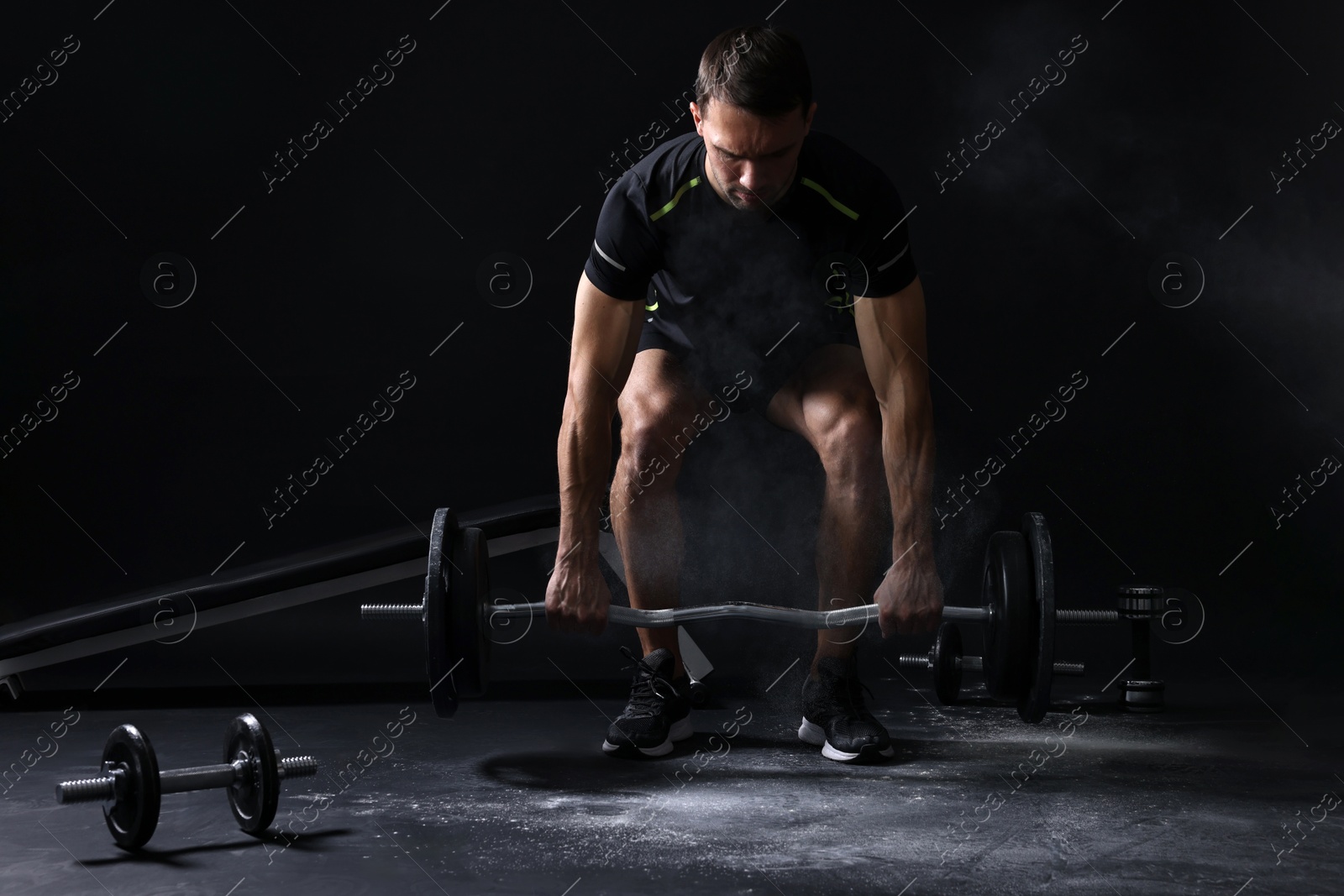 Photo of Man with talcum powder on hands training with barbell against black background