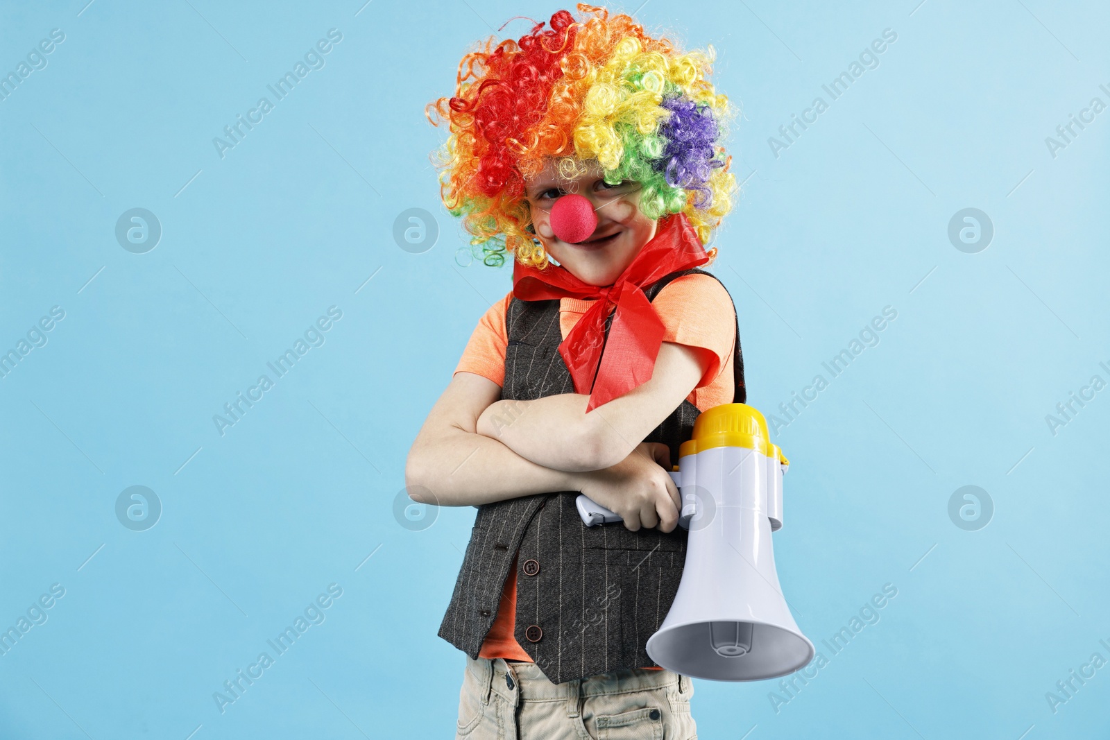 Photo of Happy little boy dressed like clown with megaphone on light blue background. Surprise party