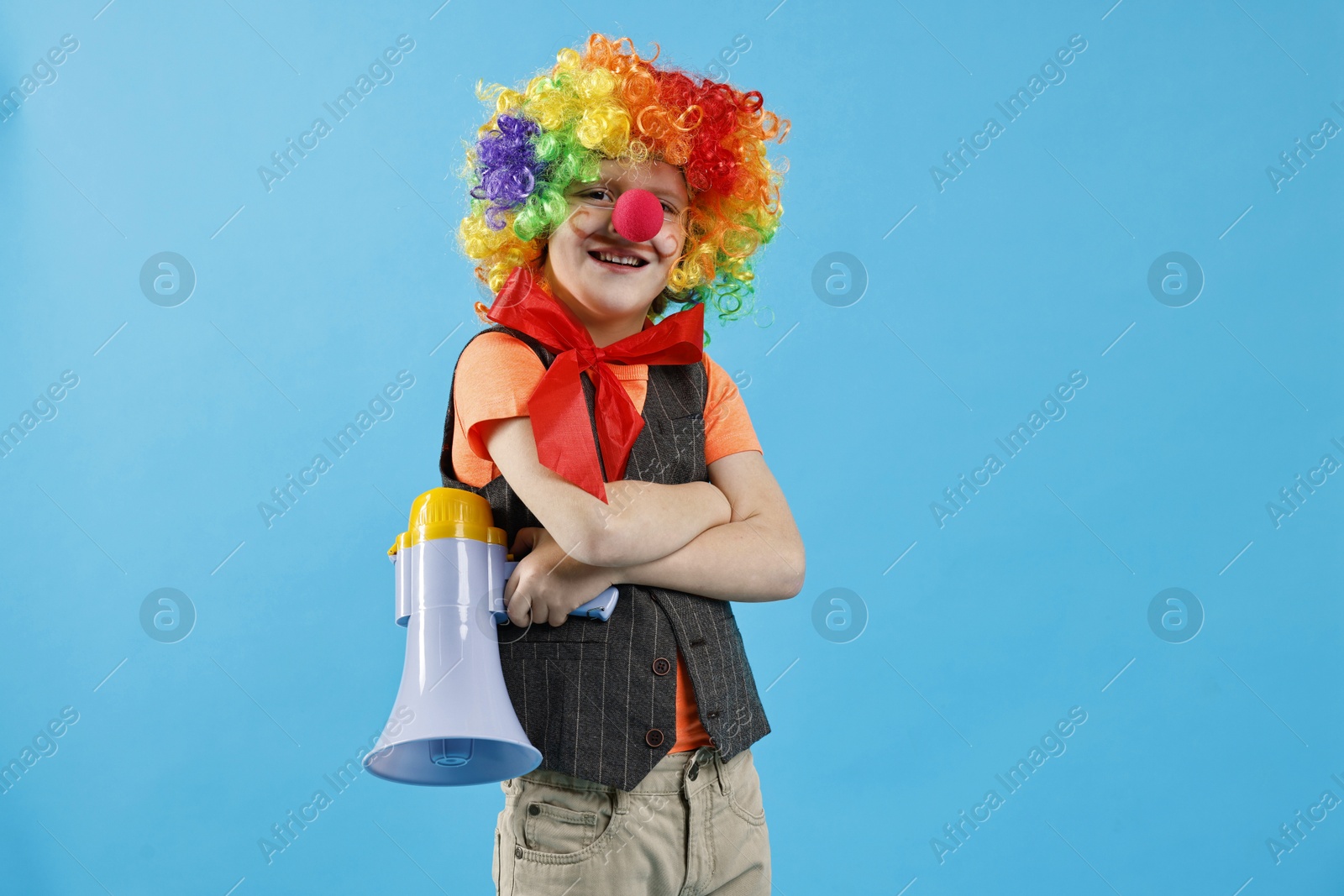 Photo of Happy little boy dressed like clown with megaphone on light blue background. Surprise party