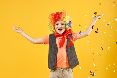 Photo of Happy little boy dressed like clown under falling confetti on orange background. Surprise party