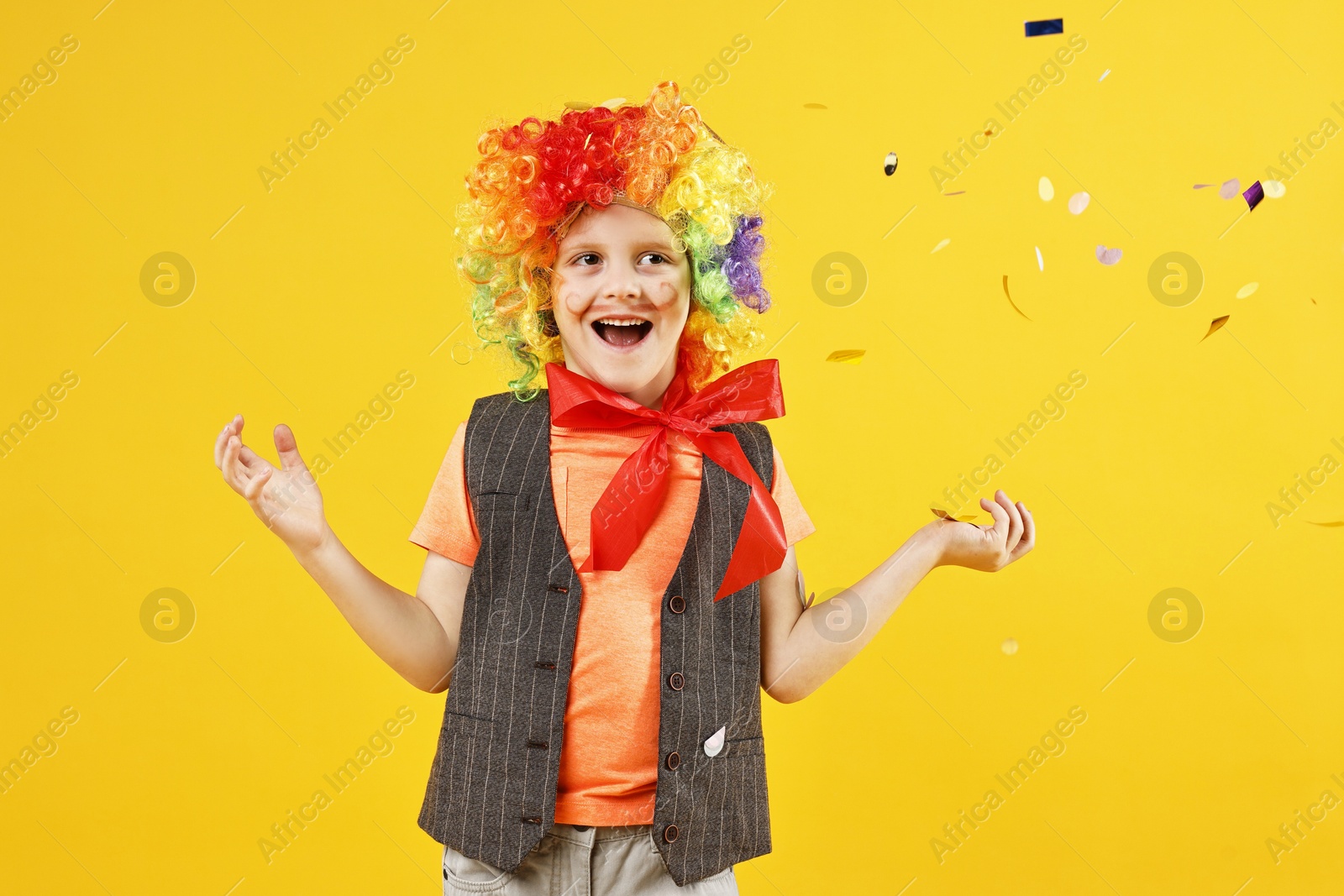 Photo of Happy little boy dressed like clown and flying confetti on orange background. Surprise party