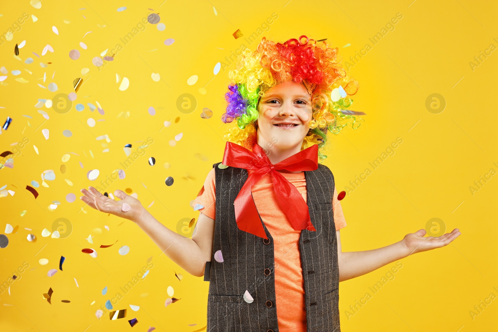 Photo of Happy little boy dressed like clown and flying confetti on orange background. Surprise party