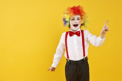 Photo of Emotional little boy dressed like clown pointing upwards on yellow background, space for text. Surprise party