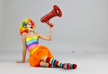 Girl dressed like clown shouting in megaphone on grey background, space for text. Surprise party