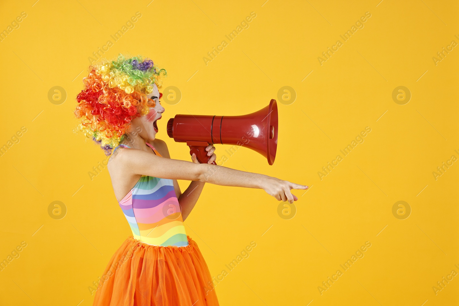 Photo of Cute girl dressed like clown shouting in megaphone and pointing at something on yellow background, space for text. Surprise party