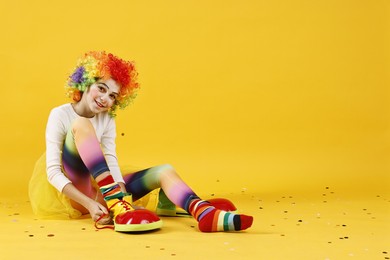 Photo of Cute girl dressed like clown and confetti on yellow background, space for text. Surprise party